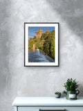 Framed photograph of a scenic landscape with a castle on a wall above a white cabinet with plants.