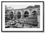 Black and white photograph of boats docked by a stone bridge over a river.