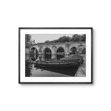 Framed black and white photograph of a boat on a river with a stone bridge in the background.