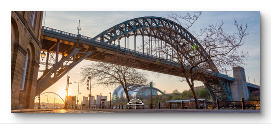 A panoramic landscape photograph of a large steel arch bridge at sunset, with bare trees in the foreground, a city street below, and a pale blue sky. The photo is presented with a white border, suggesting it is sold as a wall art print or canvas.