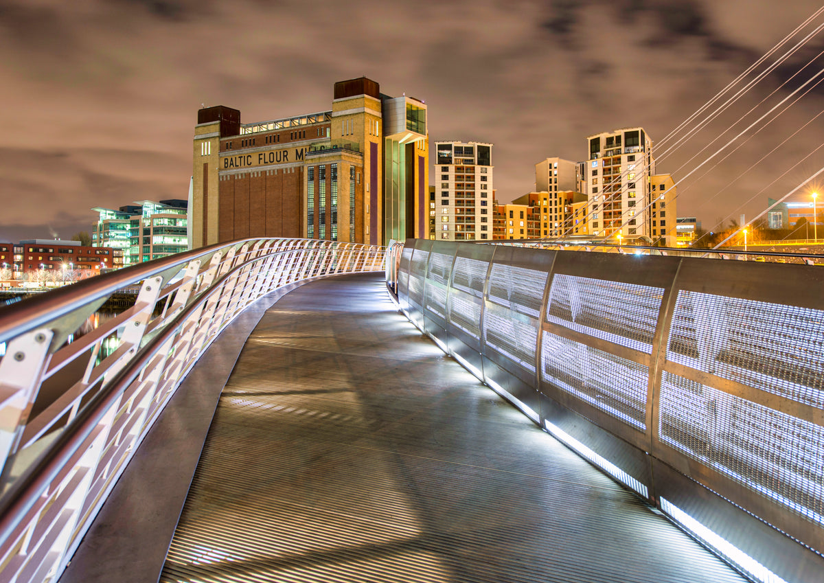 A modern elevated walkway at night with illuminated metal railings and a curved path leading toward city buildings, including a large lit building labeled 'BALTIC FLOOR MFG.'.