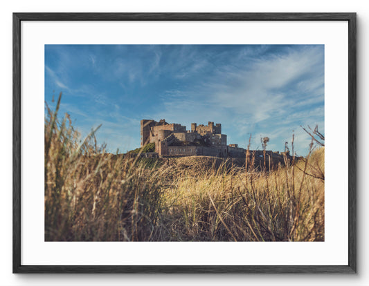 Bamburgh Castle seen through the dunes shown as a framed photographic print.
