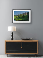 Framed landscape print of a castle on a gray wall above a wooden cabinet with a lamp.