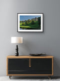 Framed landscape print of a castle on a gray wall above a wooden cabinet with a lamp.