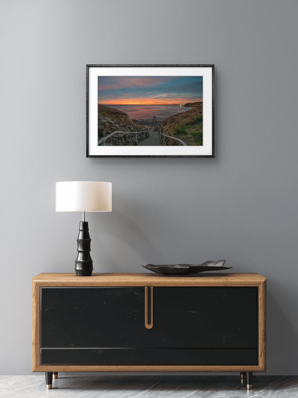 Framed artwork above a wooden cabinet with a lamp on a gray wall.