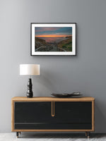 Framed artwork above a wooden cabinet with a lamp on a gray wall.