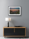 Framed artwork above a wooden cabinet with a lamp on a gray wall.