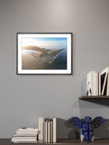 Framed artwork of a coastal scene on a gray wall with books and a decorative object below.
