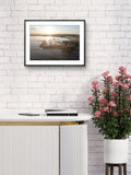 Framed artwork of a sunset over water on a white brick wall above a table with a plant.