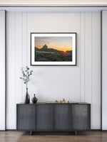 Framed artwork of a sunset over a mountain landscape on a wall above a dark piece of furniture.