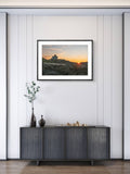 Framed artwork of a sunset over a mountain landscape on a wall above a dark piece of furniture.