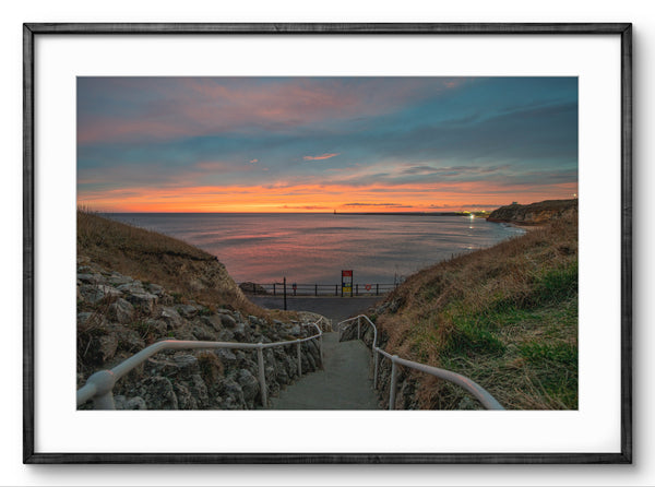 Sunset over a coastal path with a railing, framed by a black border.