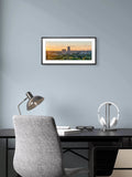 Framed artwork of a cityscape at sunset above a desk with a chair and lamp.