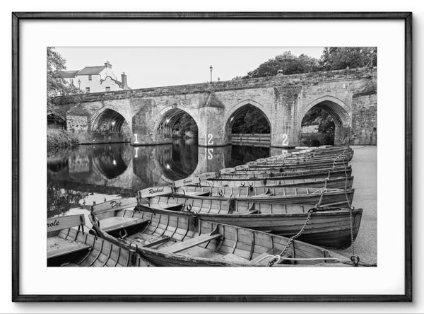 Black and white photograph of boats docked by a stone bridge over a river.