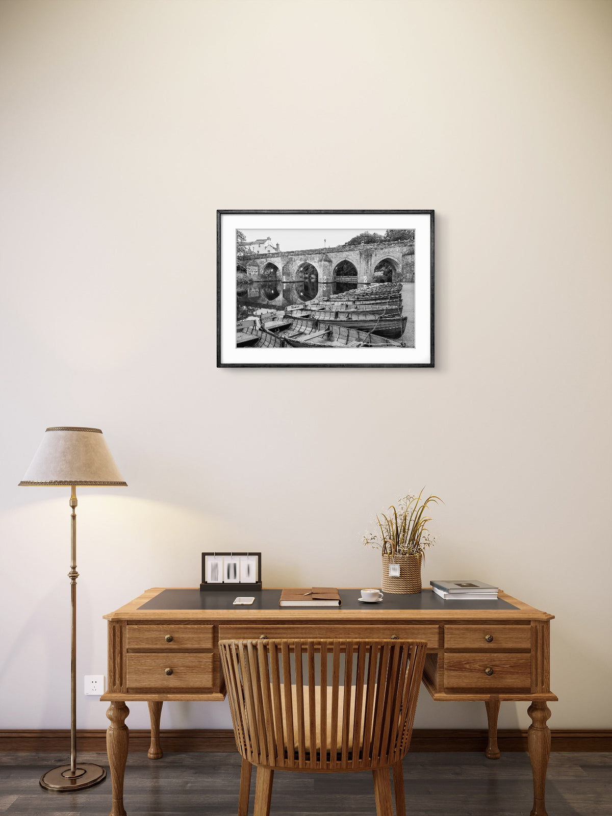 Wooden desk with chair, lamp, and framed picture on a beige wall.