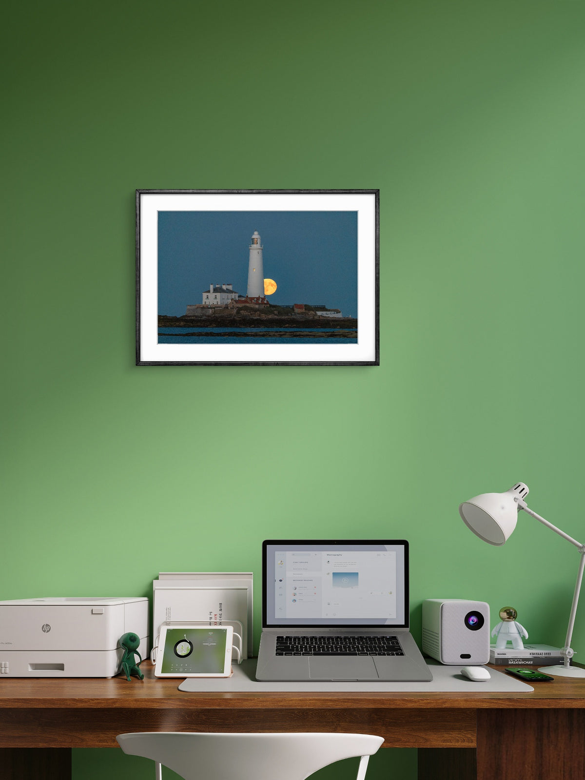 Framed picture of a lighthouse on a green wall above a desk with a laptop and printer.