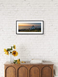 Framed landscape print of a lighthouse at sunset on a white brick wall above a wooden cabinet with sunflowers.