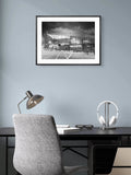 Framed black and white photo of a stadium above a desk with a chair and lamp.
