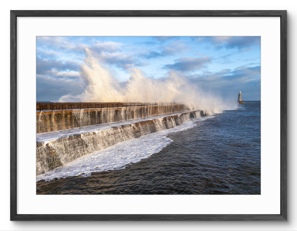 Strong waves crashing against a sea wall with a lighthouse in the distance, framed in black.