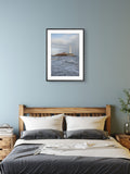 Framed artwork of a lighthouse over water above a bed in a bedroom.