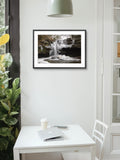 Framed photograph of a waterfall on a wall above a white desk with a laptop and coffee cup.