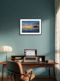Home office with a desk, chair, laptop, lamp, and framed picture on a teal wall.
