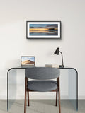 Modern office setup with a glass desk, gray chair, laptop, and framed artwork on the wall.
