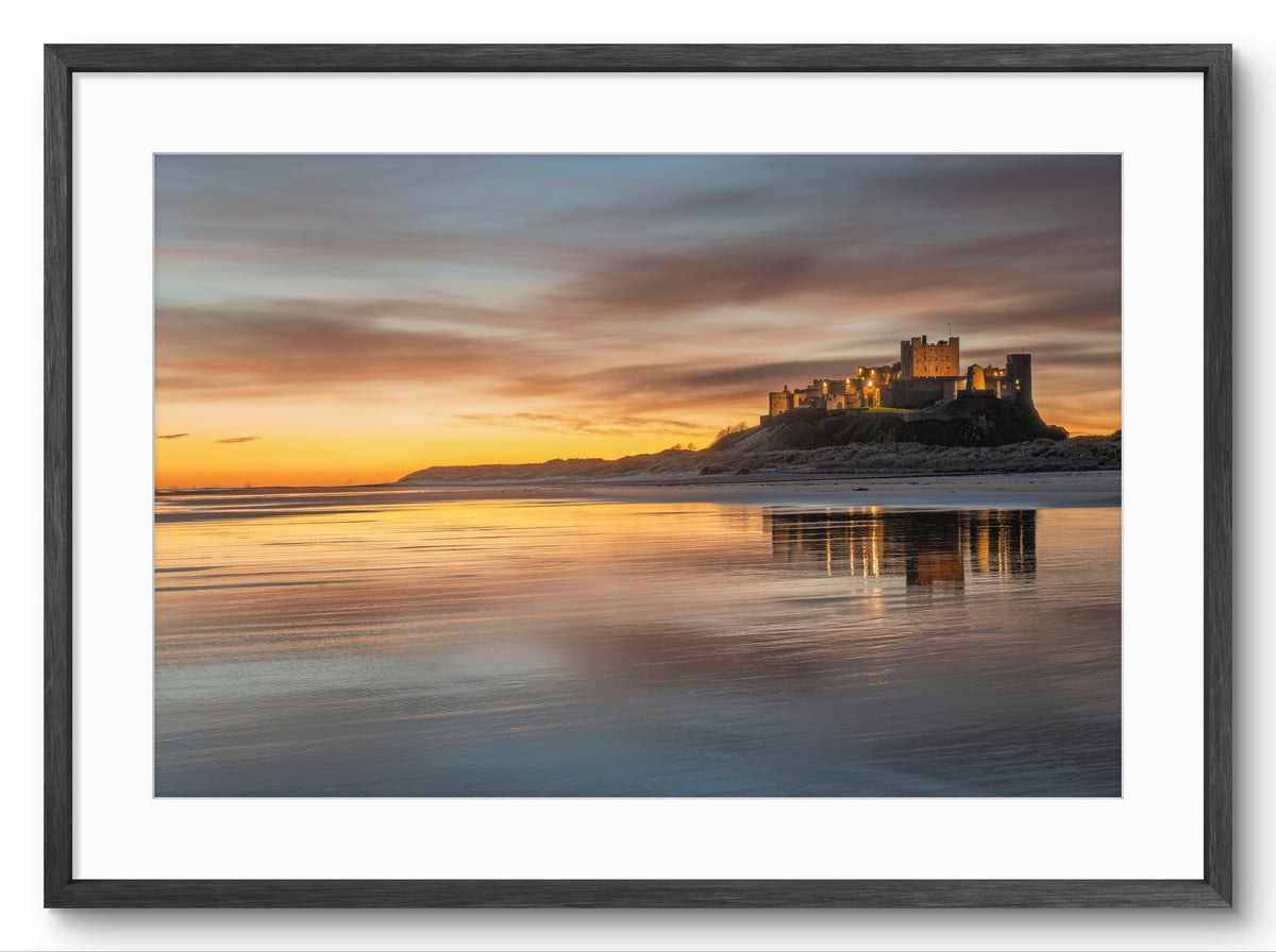 Bamburgh Castle at golden sunrise reflected on wet sand, shown as a framed photographic print.