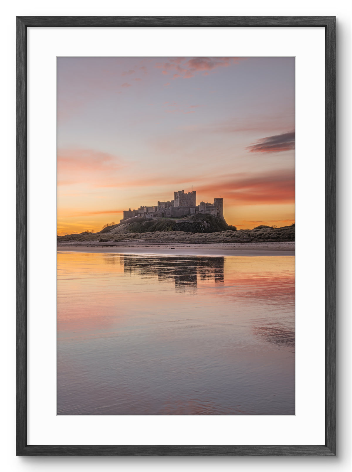 Portrait photograph of Bamburgh Castle at sunrise reflecting in wet sand with soft gold, pink and blue sky tones, Northumberland coast
