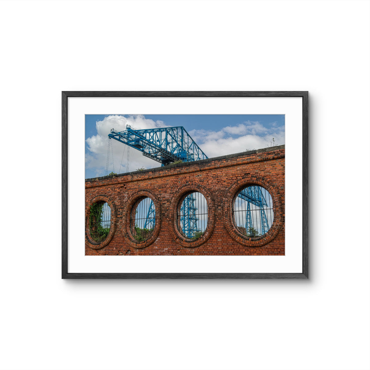 Framed photograph of a brick wall with circular windows and a blue bridge in the reflection.
