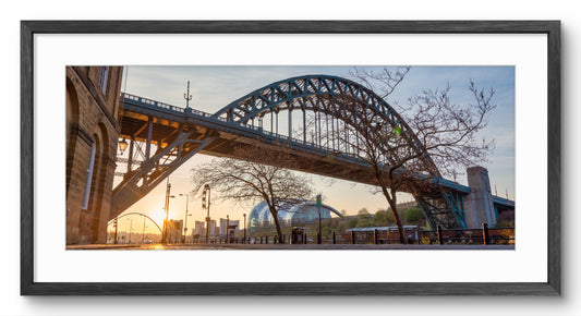 Framed panoramic wall art showing a large steel arch bridge over a city street at sunset, with bare trees in the foreground and a white mat inside a dark frame.