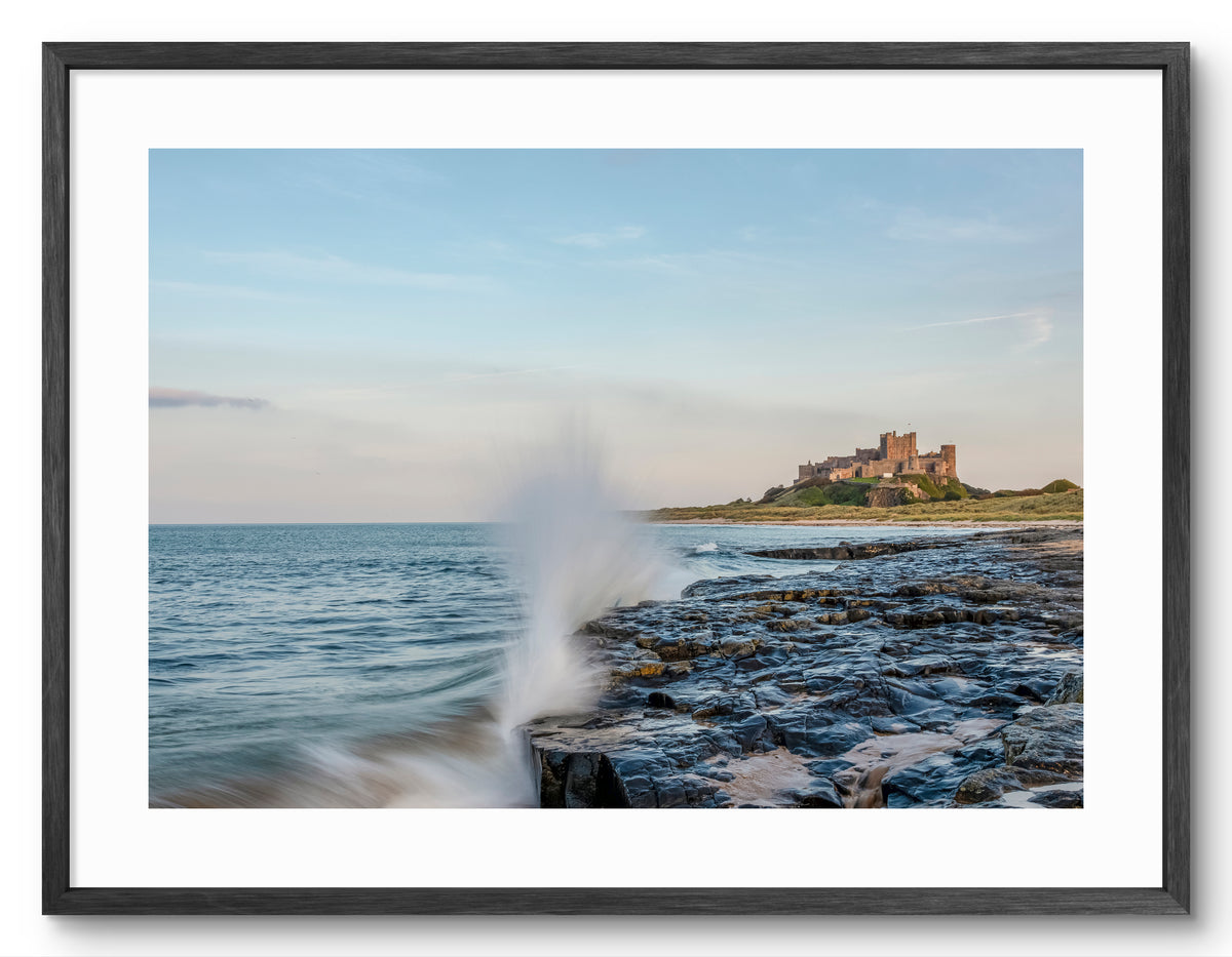 Waves crashing on rocks below Bamburgh Castle shown as a framed photographic print.