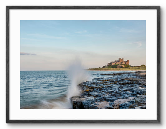 Waves crashing on rocks below Bamburgh Castle shown as a framed photographic print.