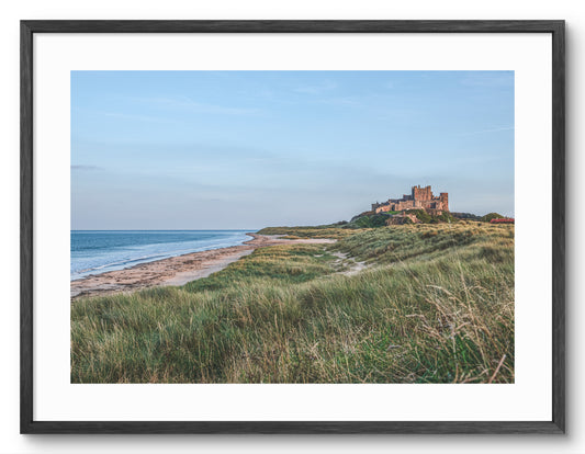 Bamburgh Castle behind grass-covered sand dunes shown as a framed photographic print.