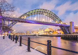 Snowy cityscape with a large arch bridge over a river at dusk.