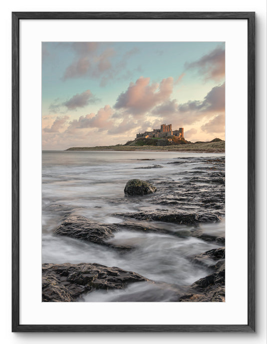 Waves on rocks at sunset near Bamburgh Castle shown as a framed photographic print.