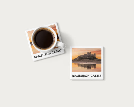 Illustrated ceramic coaster showing Bamburgh Castle reflected beneath an orange sunrise; mockup with a coffee mug.