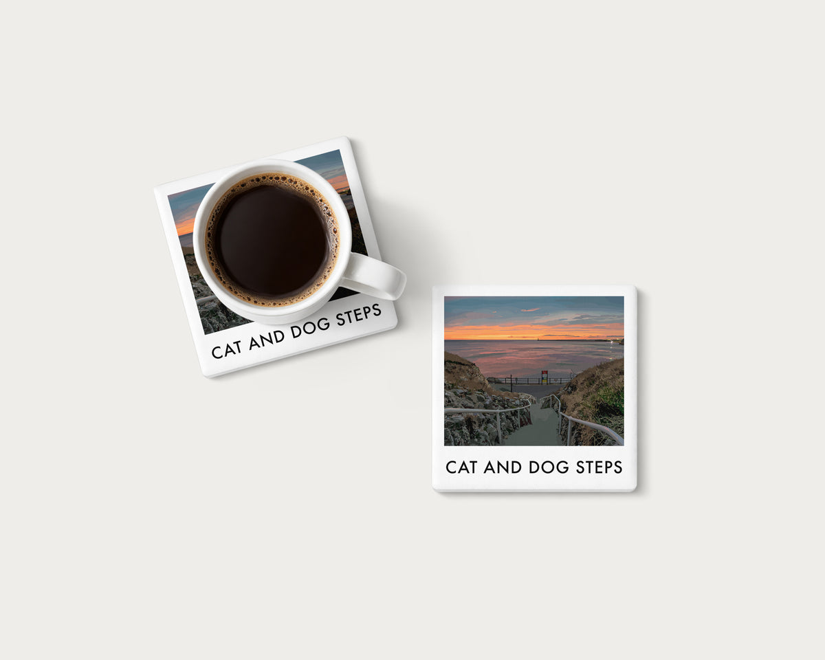 Illustrated ceramic coaster showing Cat and Dog Steps leading down towards an orange coastal sky; mockup with a coffee mug.