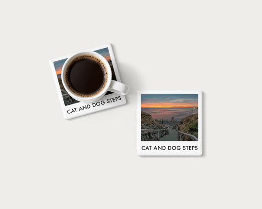 Illustrated ceramic coaster showing Cat and Dog Steps leading down towards an orange coastal sky; mockup with a coffee mug.