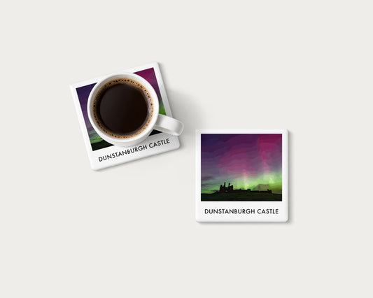 Illustrated ceramic coaster showing Dunstanburgh Castle silhouetted beneath green and purple aurora; mockup with a coffee mug.