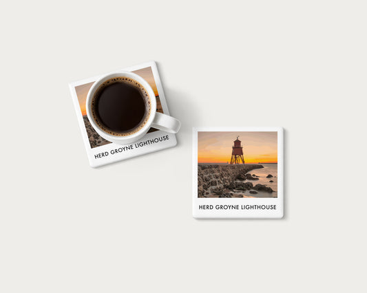 Illustrated ceramic coaster showing Herd Groyne Lighthouse above a rocky shore and orange horizon; mockup with a coffee mug.