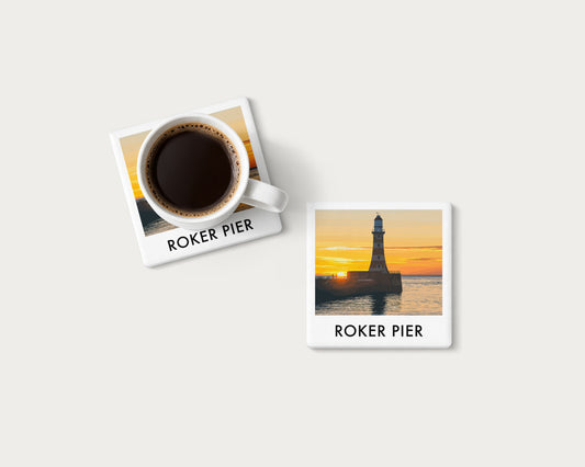 Illustrated ceramic coaster showing Roker Pier lighthouse against a golden sunrise; mockup with a coffee mug.