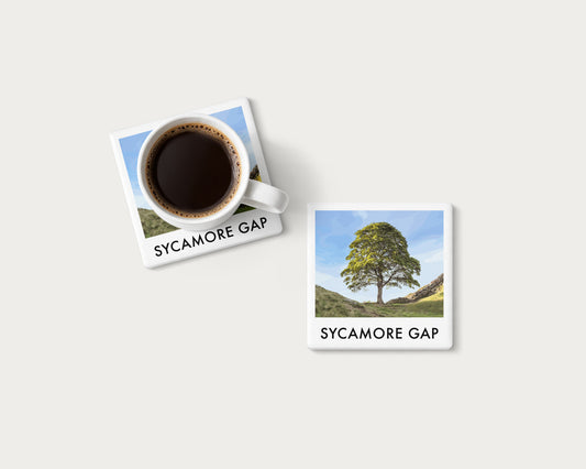 Illustrated ceramic coaster showing the Sycamore Gap tree beneath blue sky between grassy slopes; mockup with a coffee mug.