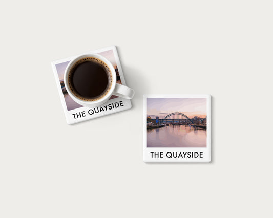 Illustrated ceramic coaster showing the Tyne Bridge above calm river reflections beneath pink sky; mockup with a coffee mug.
