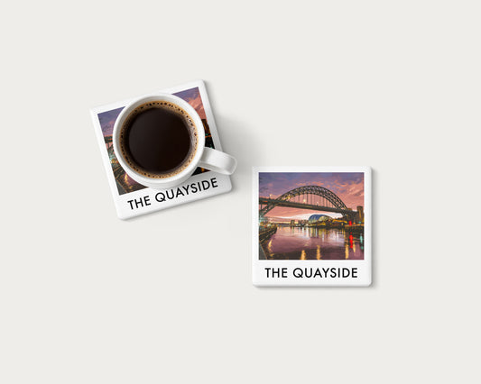 Illustrated ceramic coaster showing the Tyne Bridge and Quayside reflected beneath a colourful sunset; mockup with a coffee mug.