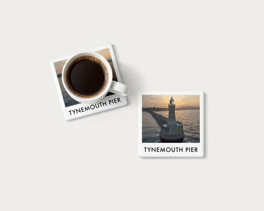 Illustrated ceramic coaster showing an aerial view of Tynemouth Pier lighthouse in golden light; mockup with a coffee mug.